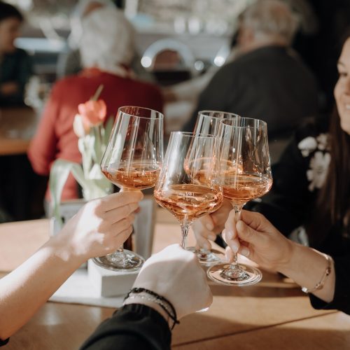 Vier Frauen stoßen mit Weingläsern an, im Hintergrund eine gesellige Feier im Café Das Eggen.