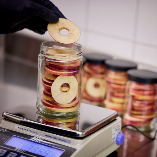 Handbefüllung von gefriergetrockneten Apfelringen in ein Glas; präzises Abwiegen und Verarbeitung in kleinen Chargen am Eggenhof.